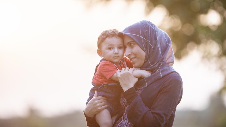 A Muslim woman is cradling her baby boy in her arms&period; They are outdoors in a park on a sunny day&period;