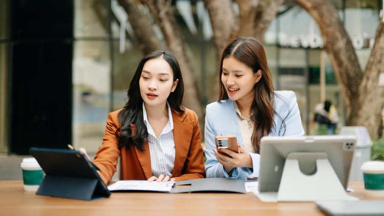 Two professional Asian businesswomen discuss strategy and documents using a digital tablet at an outdoor cafe&comma; representing modern teamwork&comma; startup life&comma; and female