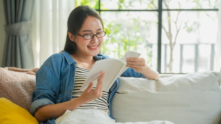 Relaxed Asian woman with glasses reading book on sofa at home&comma; smiling while enjoying leisure time in cozy living room&comma; mindfulness and slow living lifestyle concept