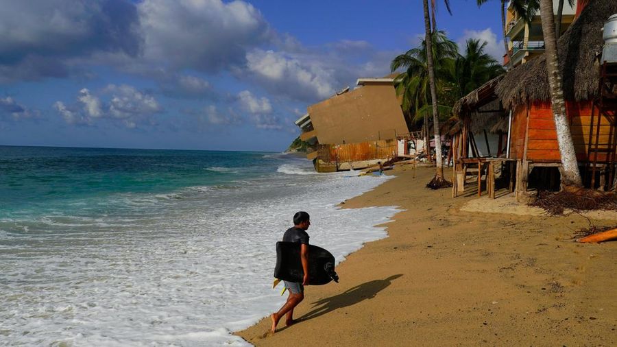 Seseorang membawa papan seluncur air (bodyboard) di pantai di depan sebuah bangunan kondominium yang rusak, setelah serangkaian badai menghantam pesisir Pasifik Meksiko, di San Francisco, Bahia de Banderas, negara bagian Nayarit, Meksiko, 21 Oktober 2026.