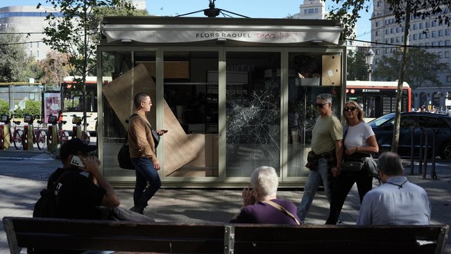 People walk past a flower kiosk damaged the previous night during riots, in Barcelona on October 25, 2026. Police made 128 arrests overnight in Barcelona for acts of "vandalism", damage to public property" and "looting", the regional Catalan police s